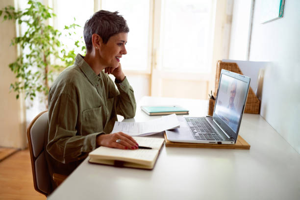 Donna con capelli corti sorridente durante una videochiamata di lavoro, seduta alla scrivania con laptop, quaderno e documenti, in un ambiente luminoso e accogliente.