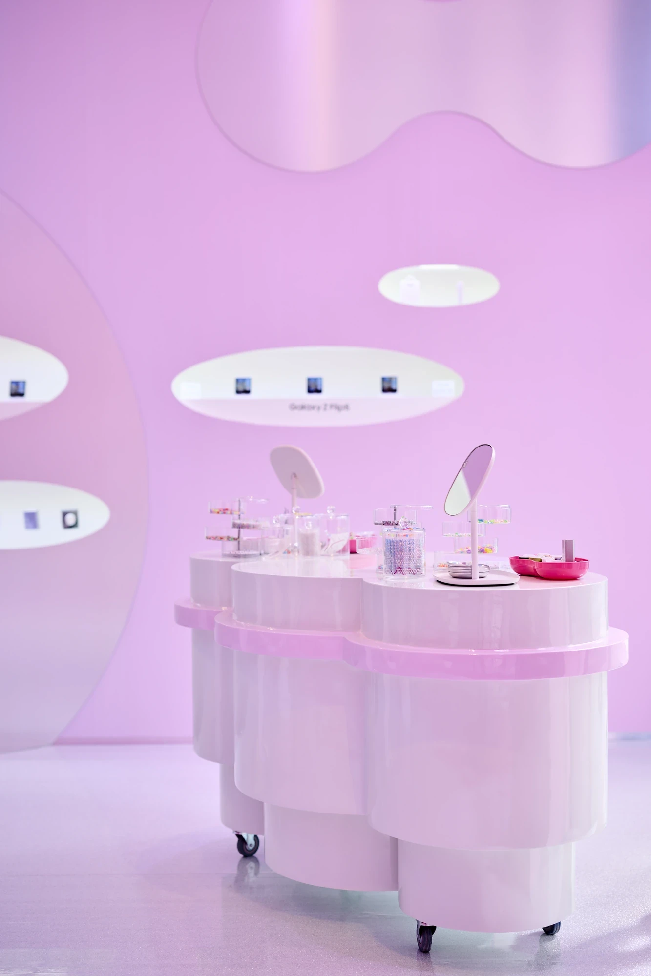 Samsung Galaxy Z Flip6 jewellery workshop area with glossy white pill-shaped counter, beadwork supplies and product display niches set into a pink iridescent wall.