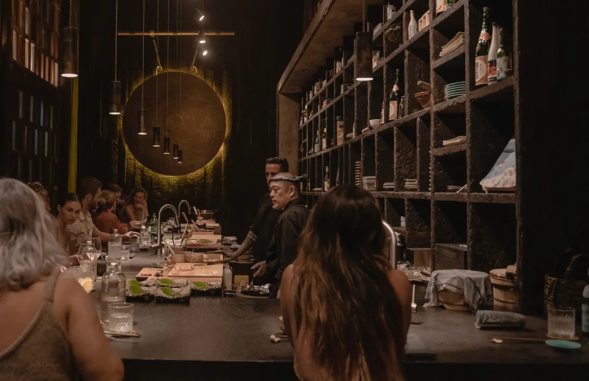 Vista del bar en el restaurante japonés Kuu en Nômade Tulum, México, con mostrador y asientos para los huéspedes, con personas sentadas y ordenando en el bar, chefs de comida japonesa atendiendo, y una atmósfera bien iluminada y decorada.