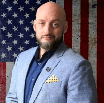 A man with a bald head and beard, wearing a light grey blazer over a dark blue shirt, stands confidently in front of an American flag backdrop.
