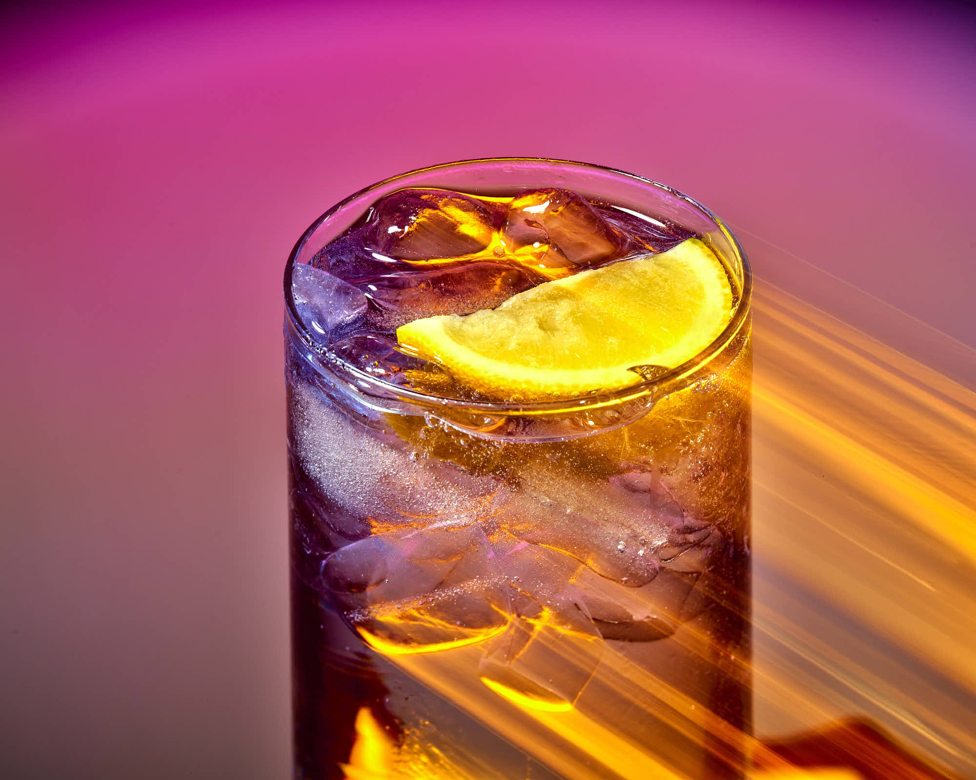 A tall glass of dark drink with ice and a slice of orange, set against a vibrant purple background.