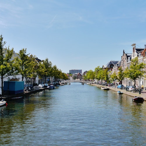 A serene canal flanked by trees and buildings on both sides, with boats tied along the edges under a clear blue sky.