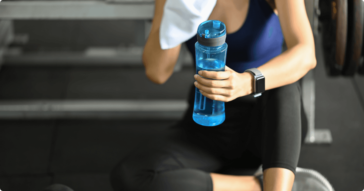  A person sitting on the floor in a gym, holding a blue water bottle in one hand. They appear to be taking a break, with a towel in the other hand and wearing athletic attire, including a fitness tracker on their wrist.