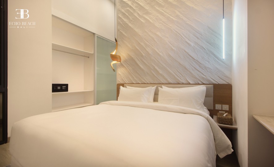 A modern bedroom at Echo Beach Suite in Canggu, Bali, featuring a comfortable white bed, a textured accent wall, warm ambient lighting, and an open wardrobe, offering a relaxing stay near Echo Beach.