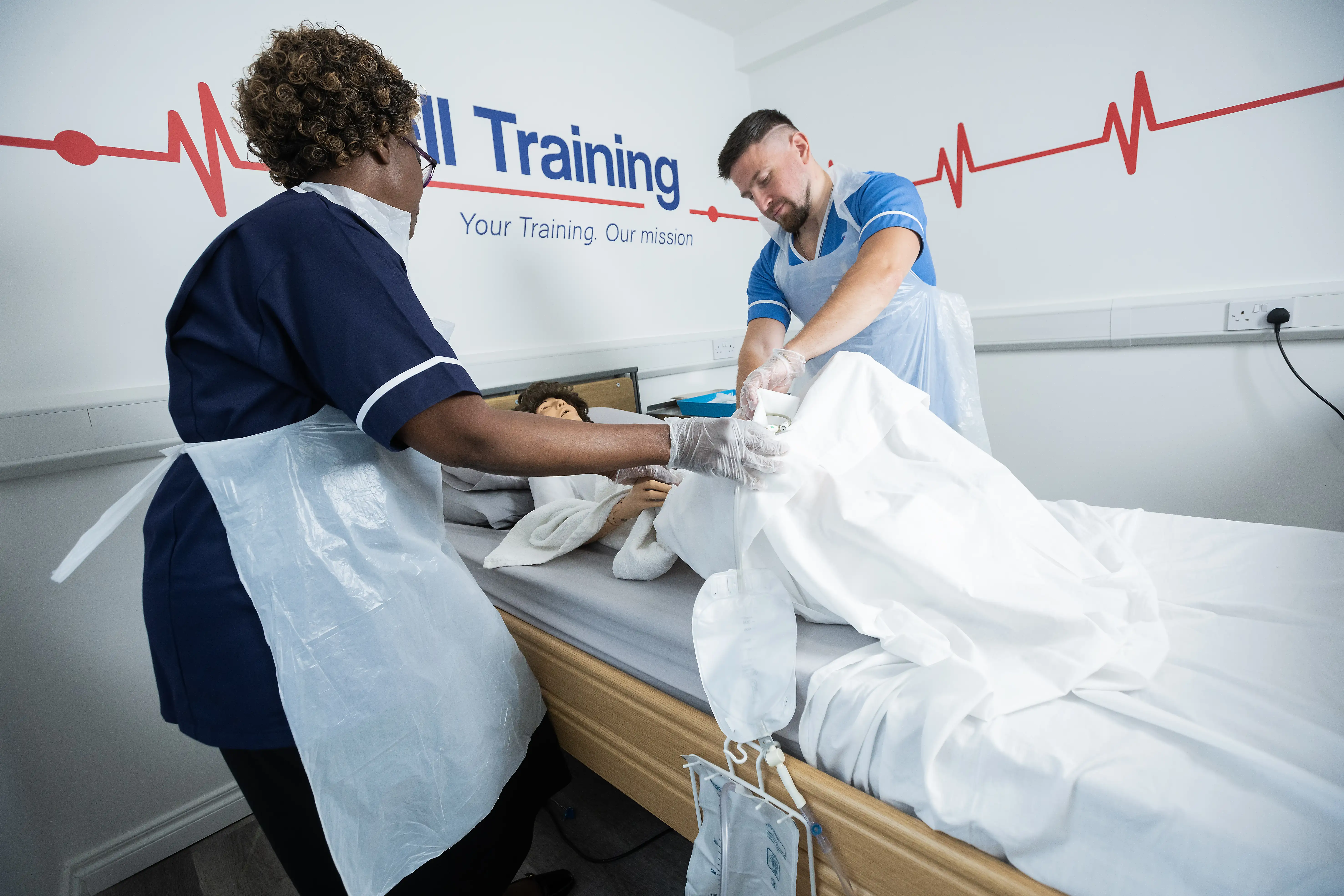 Two healthcare professionals wearing aprons and gloves demonstrating catheter care on a training mannequin in a clinical skills classroom.