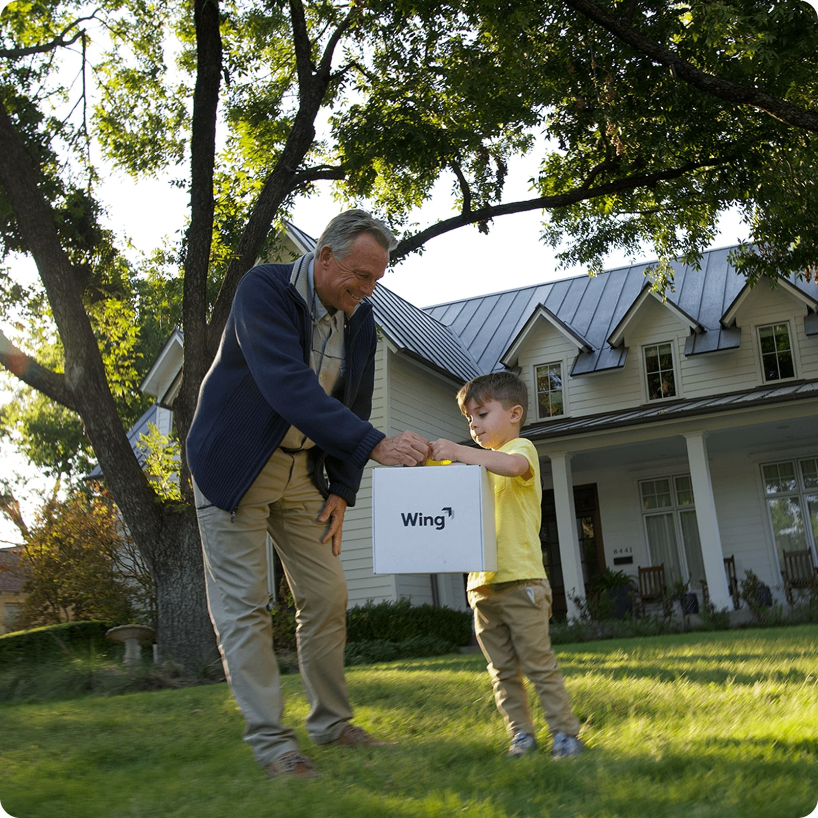 A grandfather and child stand on a lush green lawn in front of a large, charming house with tall trees, as they receive a drone delivery package marked "Wing".