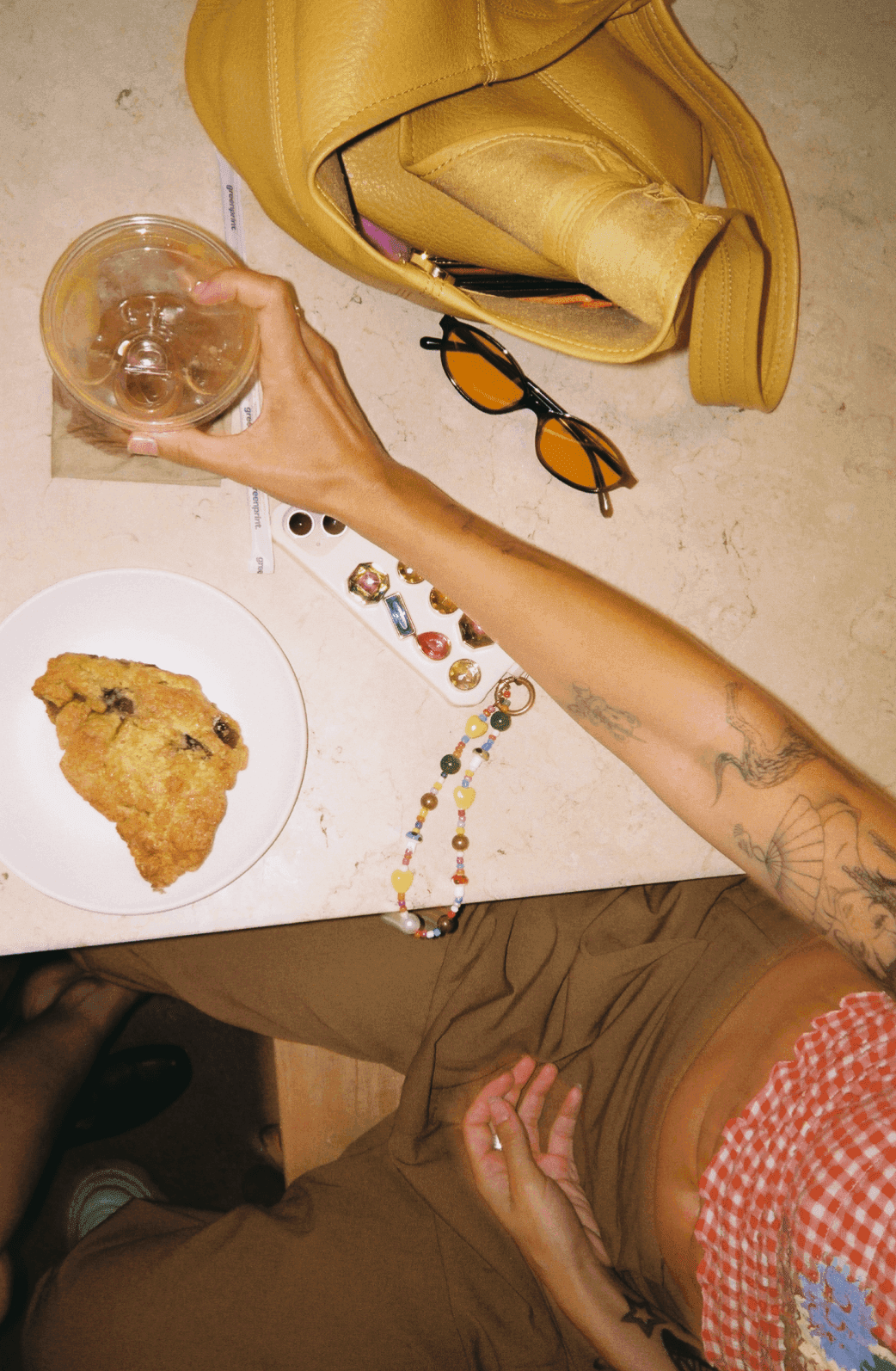 a table shot from above with sunglasses, a yellow purse, a scone, a beaded phone chain, and a hand reaching in to grab an iced coffee