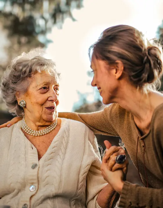 A younger woman leaning in warmly toward an older woman, holding her hand and sharing a gentle, connected moment together outdoors.