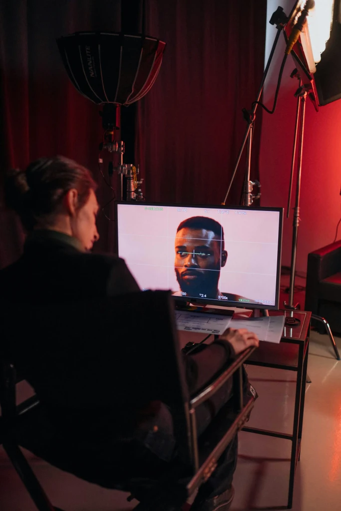 A person views a monitor showing a man's face on a film set with red studio lighting and production equipment.