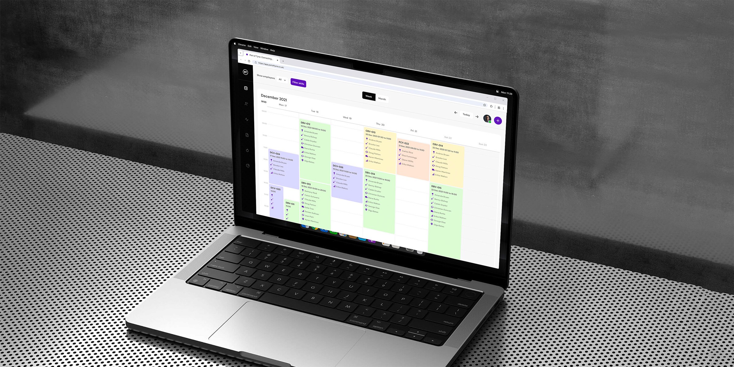 A sleek laptop with a high-resolution display shows a detailed calendar dashboard interface, featuring various color-coded entries, positioned on a modern, perforated metal desk.