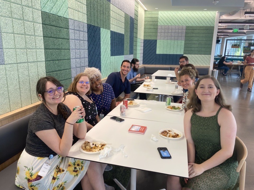 A group of eight smiling people seated at a long table, enjoying lunch together in a bright, modern dining space with green and blue geometric wall panels. Plates of food, drinks, and phones are on the table. This is the Post-Acute Care Ed Tech team during an on-site lunch.
