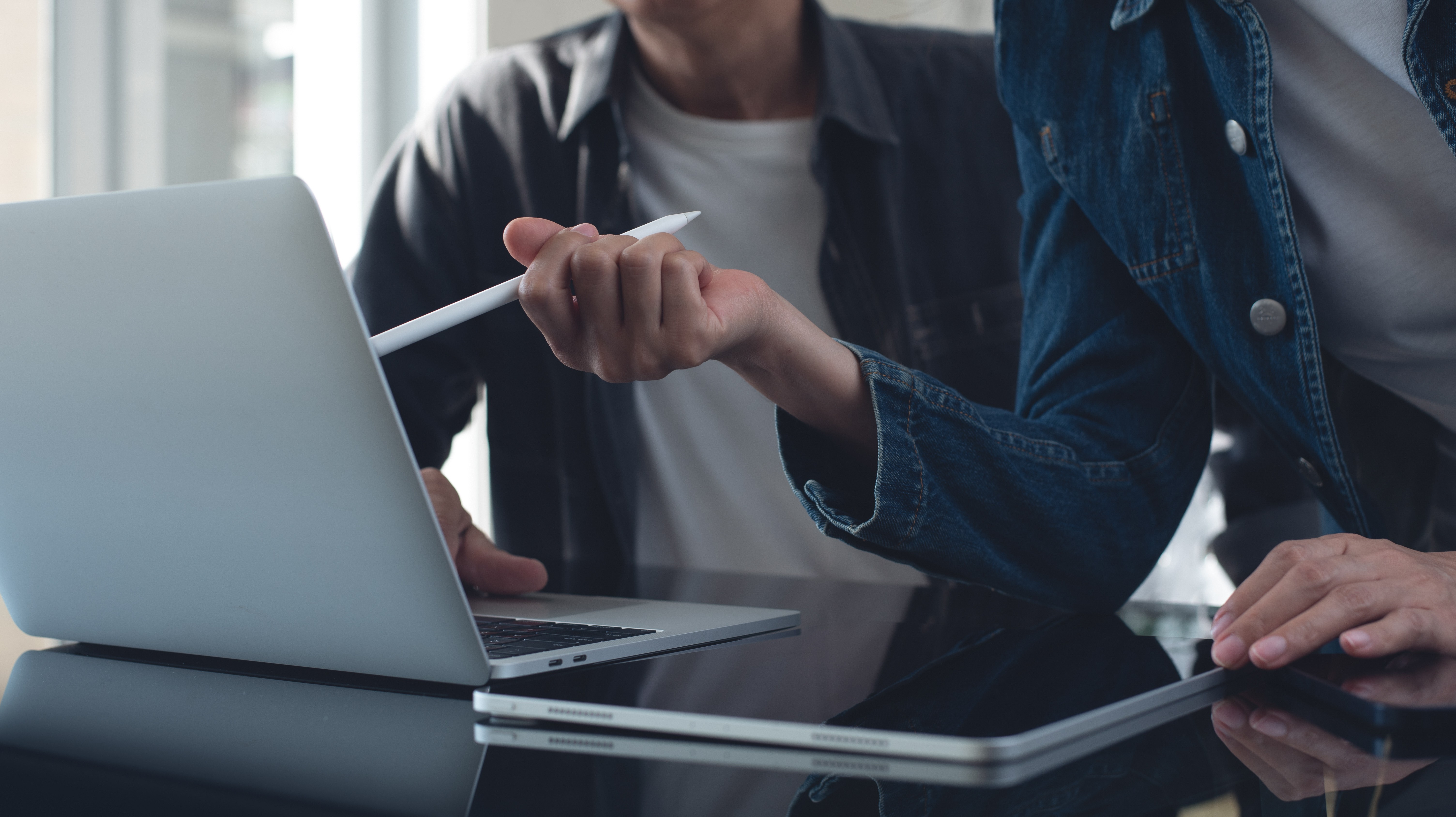 Mesa com computador e tablet. tem duas pessoas à frente conversando sobre algo do computador