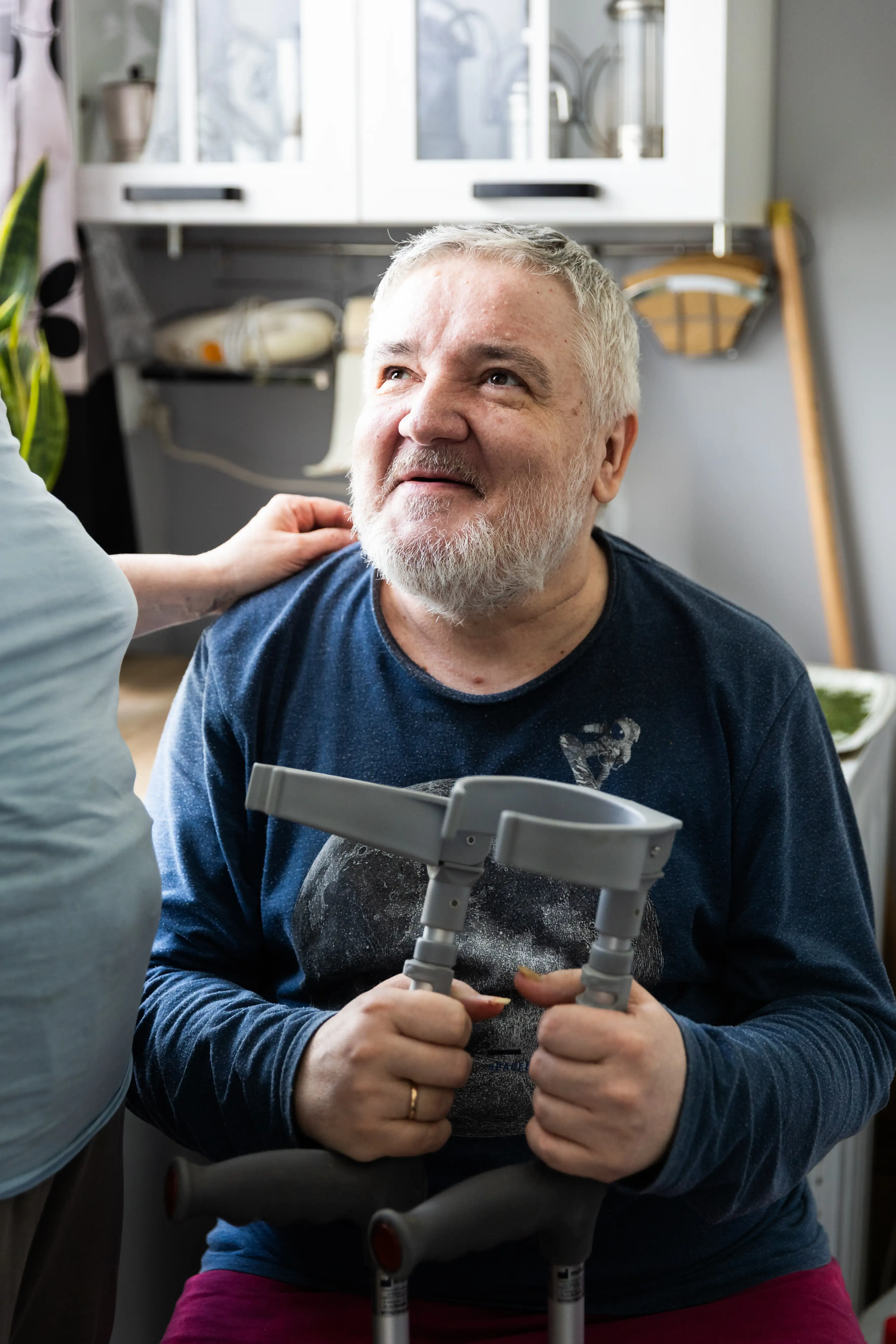 Starszy, uśmiechnięty mężczyzna siedzi na krzesle, opierając ręce na kolanach. Obok niego stoi kobieta; w tle widać mieszkanie.