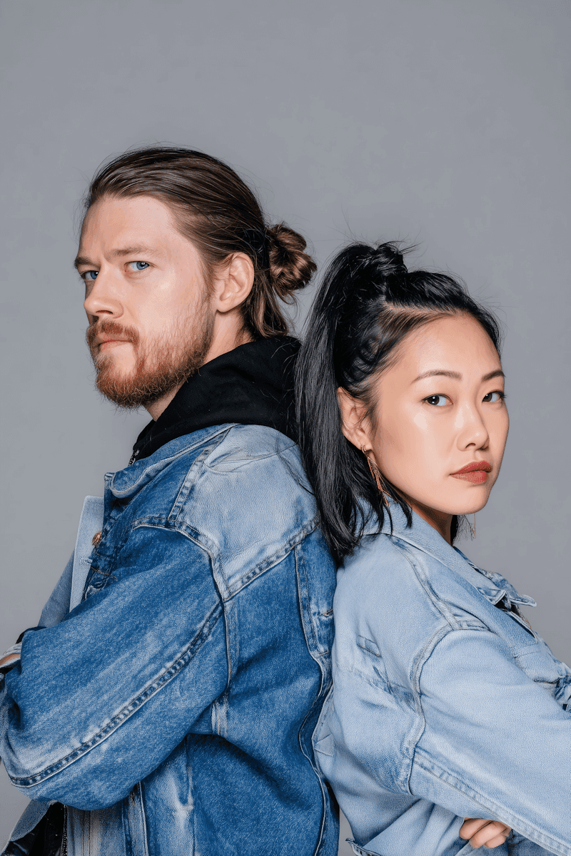 A man with a beard and a woman with long black hair stand back-to-back, both wearing denim jackets against a plain gray background.