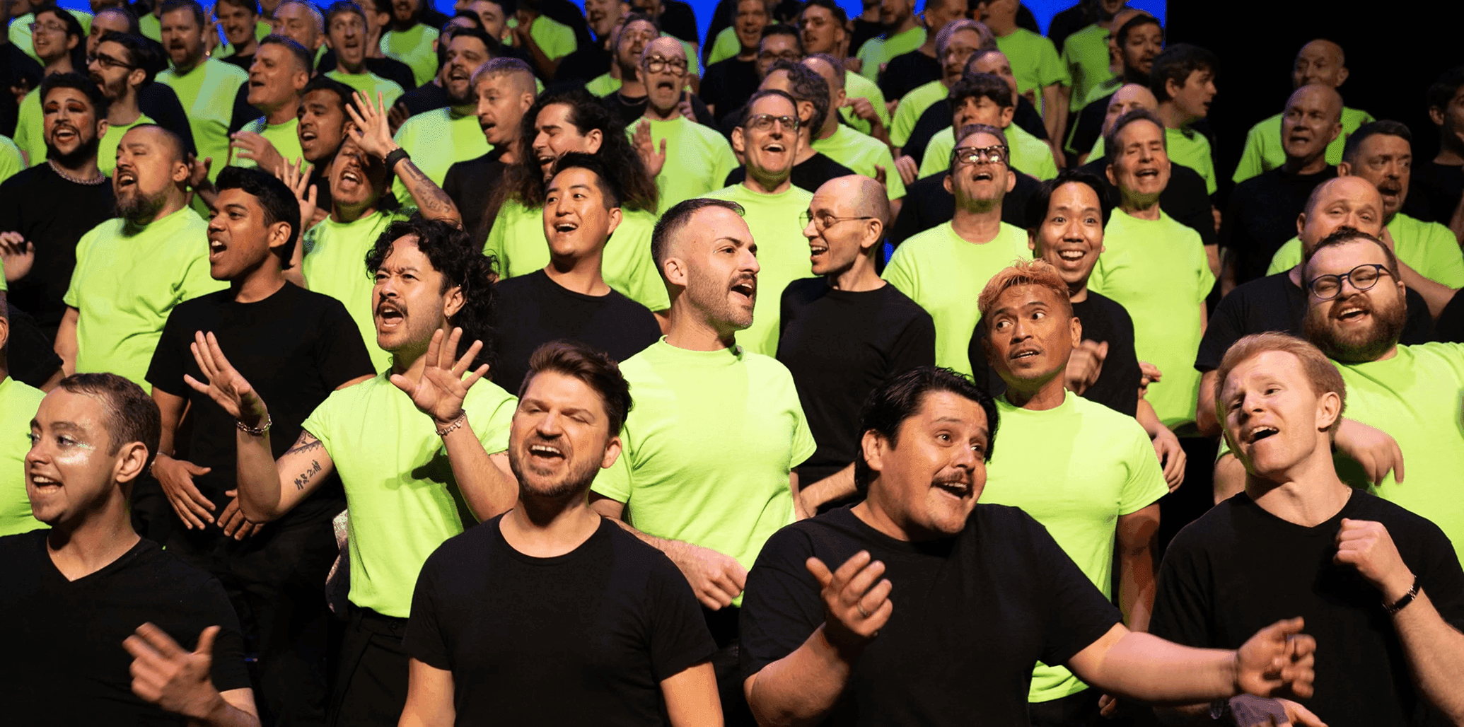 A large choir performs energetically on stage, wearing alternating neon green and black t-shirts