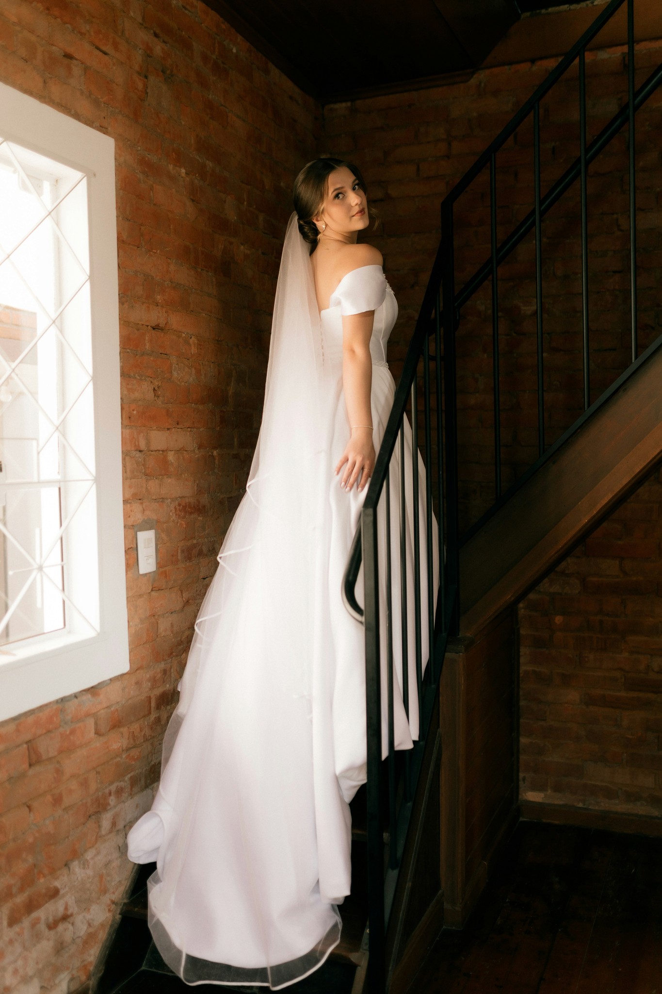 Bride in an off-the-shoulder gown and cathedral veil posing on an iron staircase against an exposed brick wall