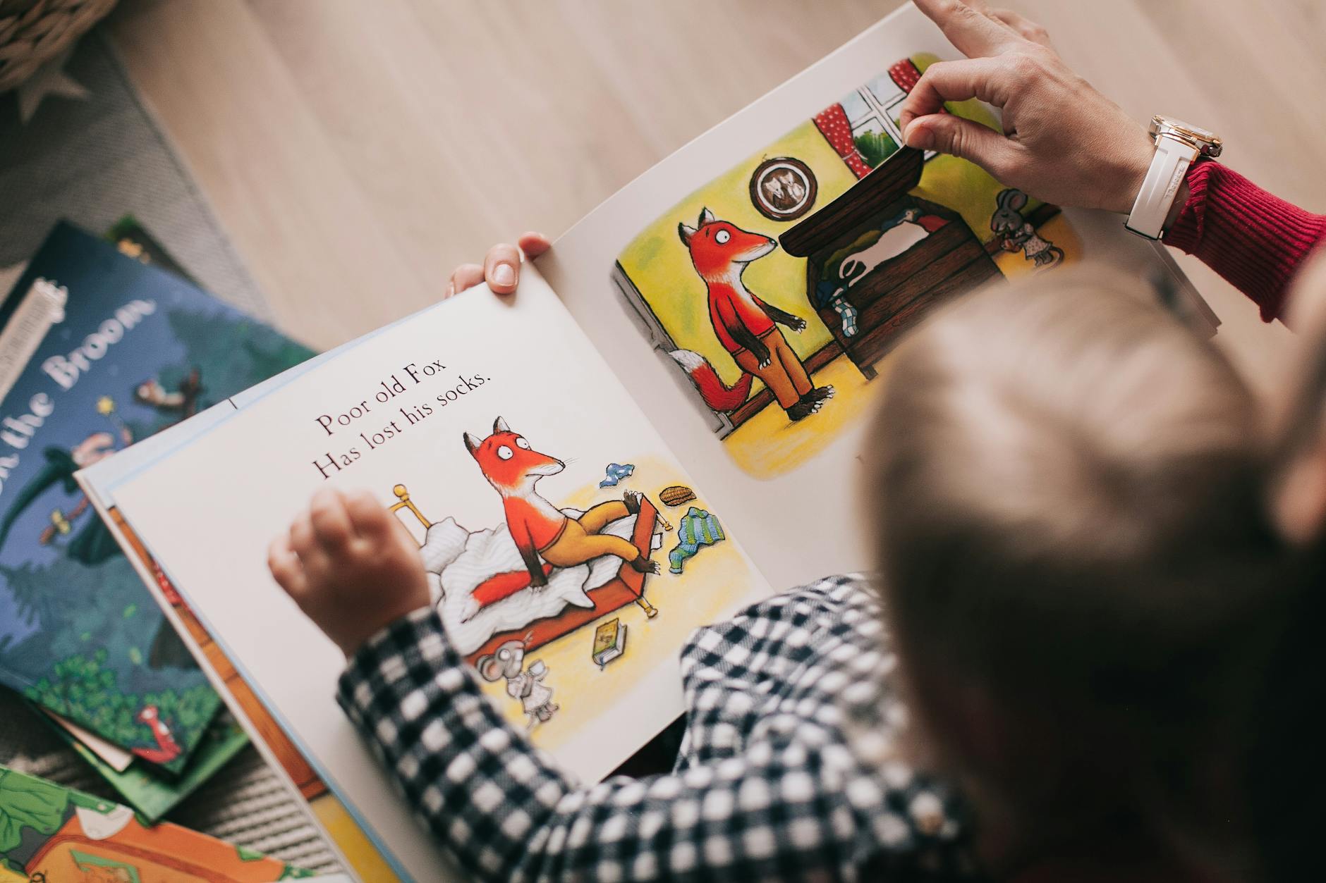 A young child sits on a cozy classroom rug holding open pre reader books while pointing at words.