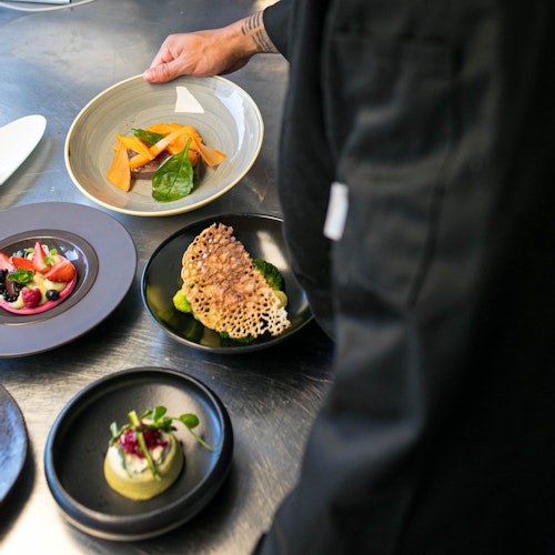 A chef in a black uniform arranges three gourmet dishes with vegetables, garnishes, and intricate plating on a metal surface.