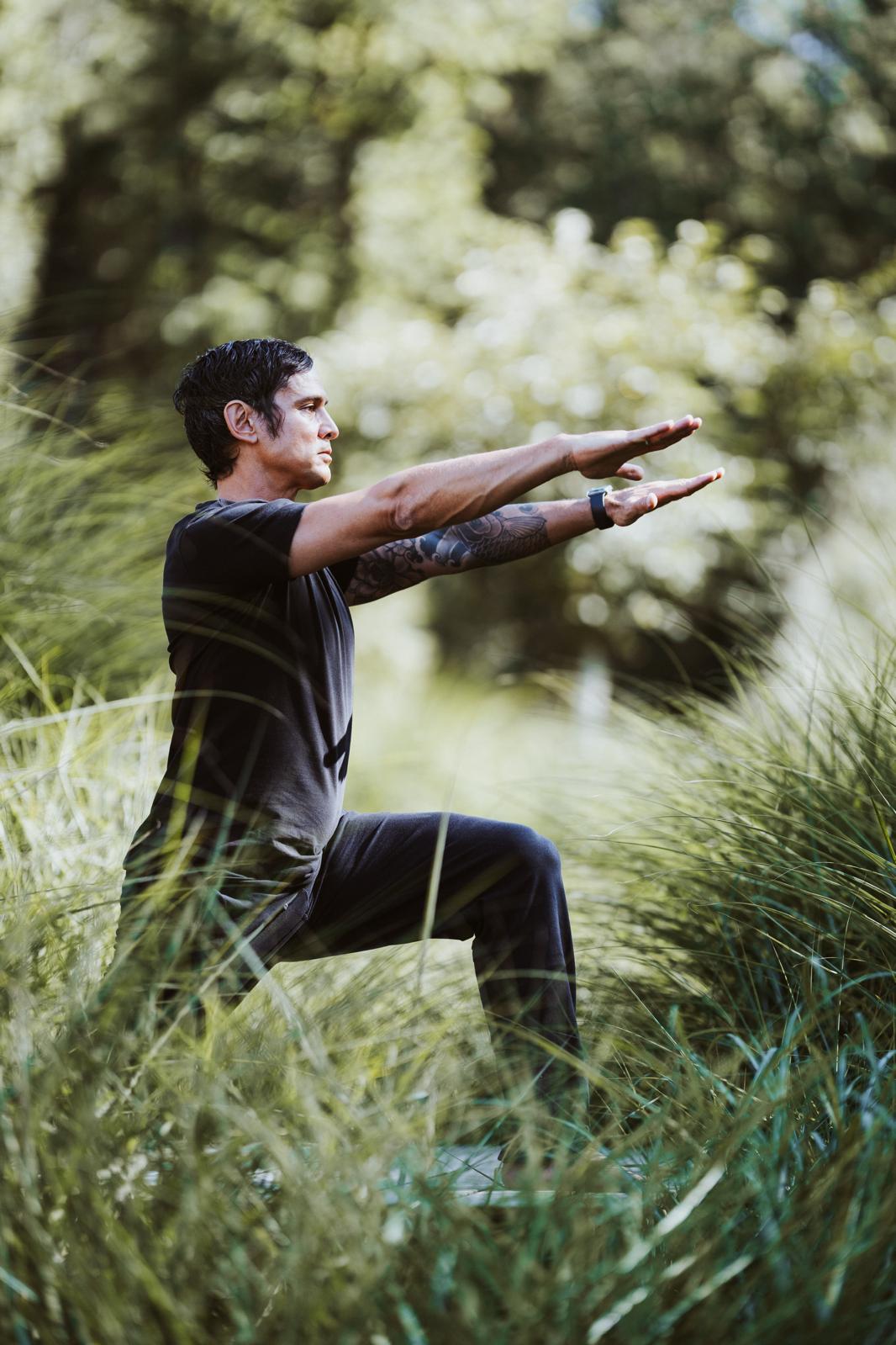A person is practicing yoga outdoors surrounded by tall, green grass and trees, performing a balance pose with arms outstretched and one knee bent, reflecting focus and tranquility in nature.