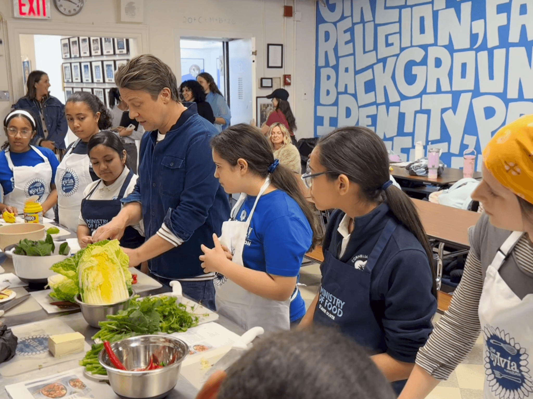 Two teachers demonstrate how to raise their hands and wave to their young students, some of whom follow along. 