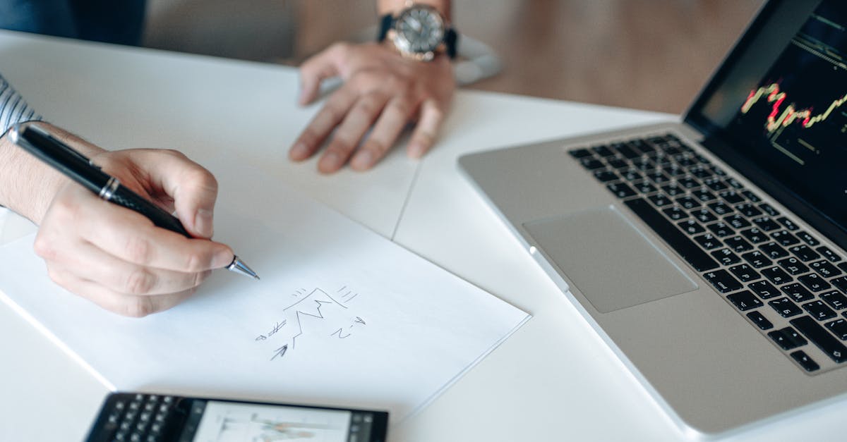 Close-up of hands drawing stock trends on paper with laptop and smartphone on desk.