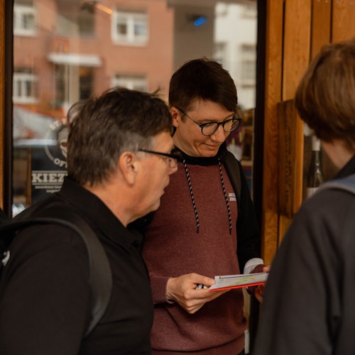 Three people stand outside a shop; one man reads a pamphlet while the others look on.