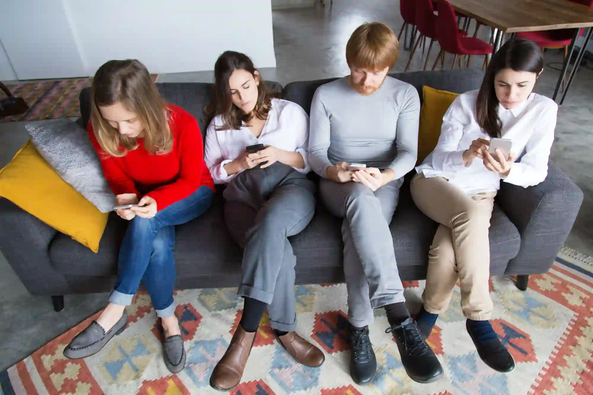 Four young adults sit together on a sofa, each distracted and focused entirely on their individual smartphone screens.