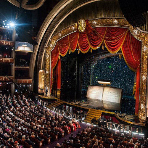 Elegant theater filled with seated audience; stage with open book prop, red curtains, and ornate gold arch.