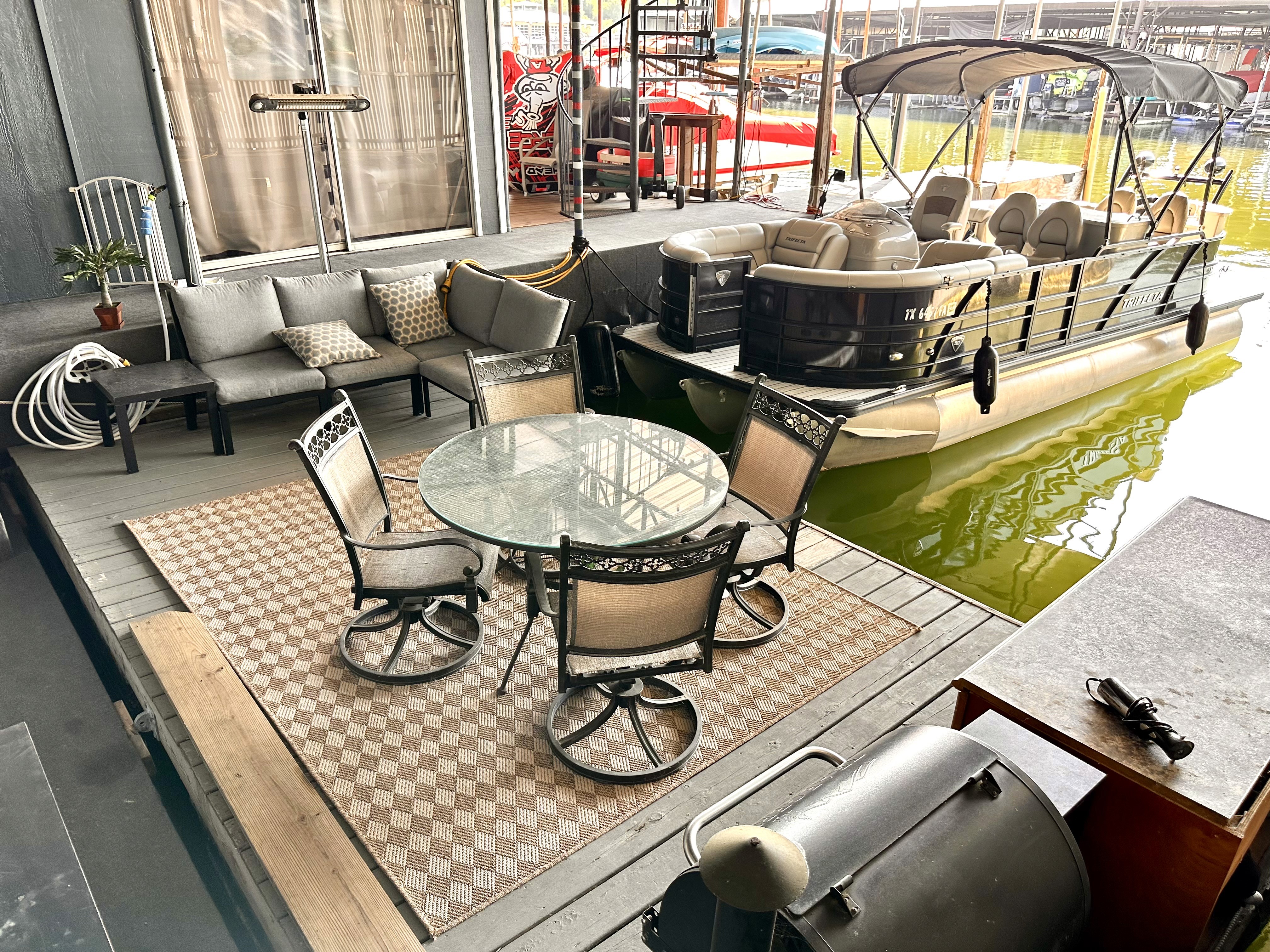 A cozy patio setup with a glass-top table and four chairs on a checkered rug, adjacent to a pontoon boat docked on a greenish-yellow waterway.