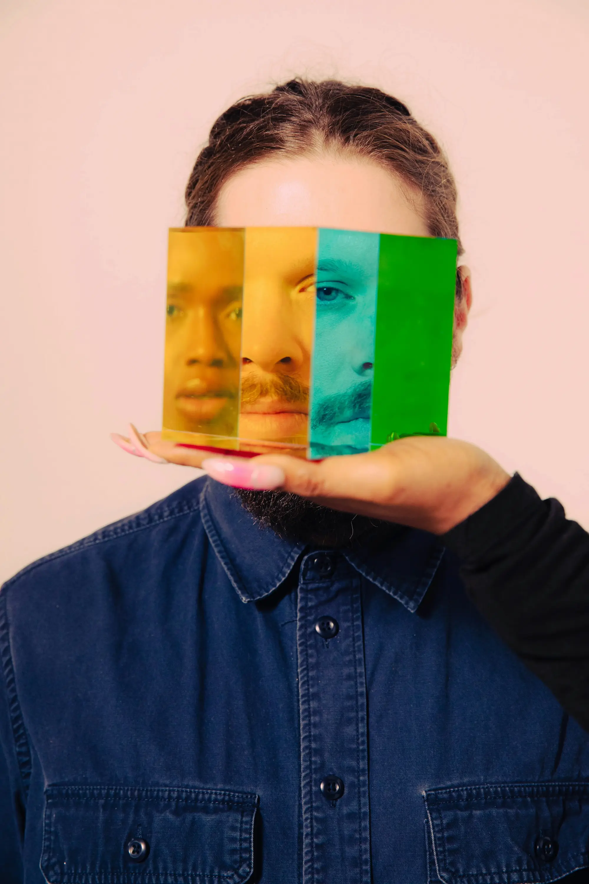 A person with long hair and a beard looks through a set of colorful translucent panels held in front of their face, creating multiple fragmented reflections in yellow, blue, and green. A hand with long pink nails supports the panels. The creative portrait symbolizes identity exploration, self-expression, and fluidity within the queer community.