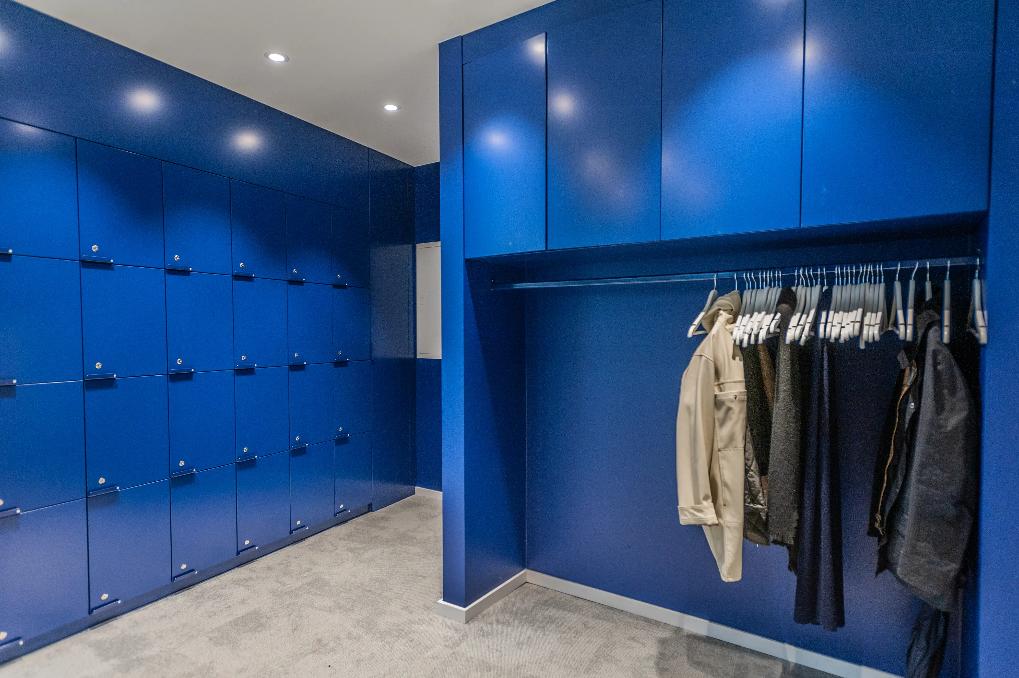 Royal-blue wardrobe room at ZENLOOP with floor-to-ceiling locker walls, an integrated hanging rail with jackets on white hangers and grey carpet.