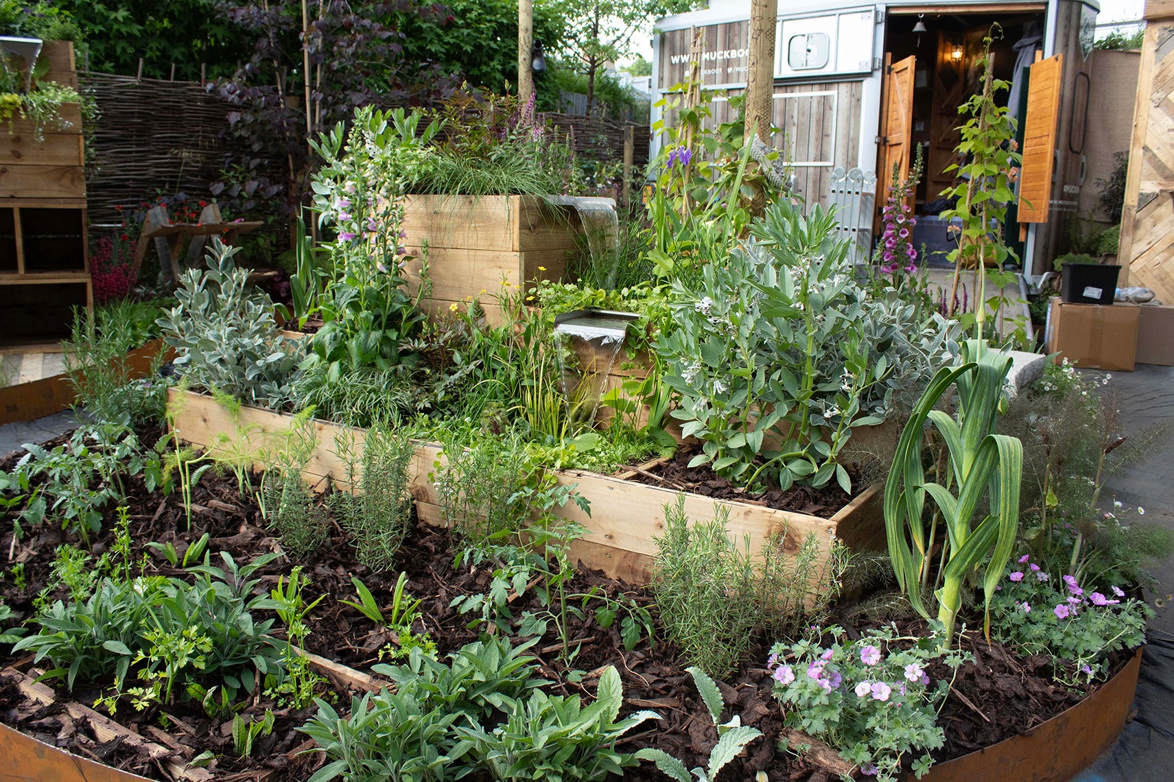 Lush garden with various plants, including herbs and flowers, surrounded by wooden planters and a rustic entrance.