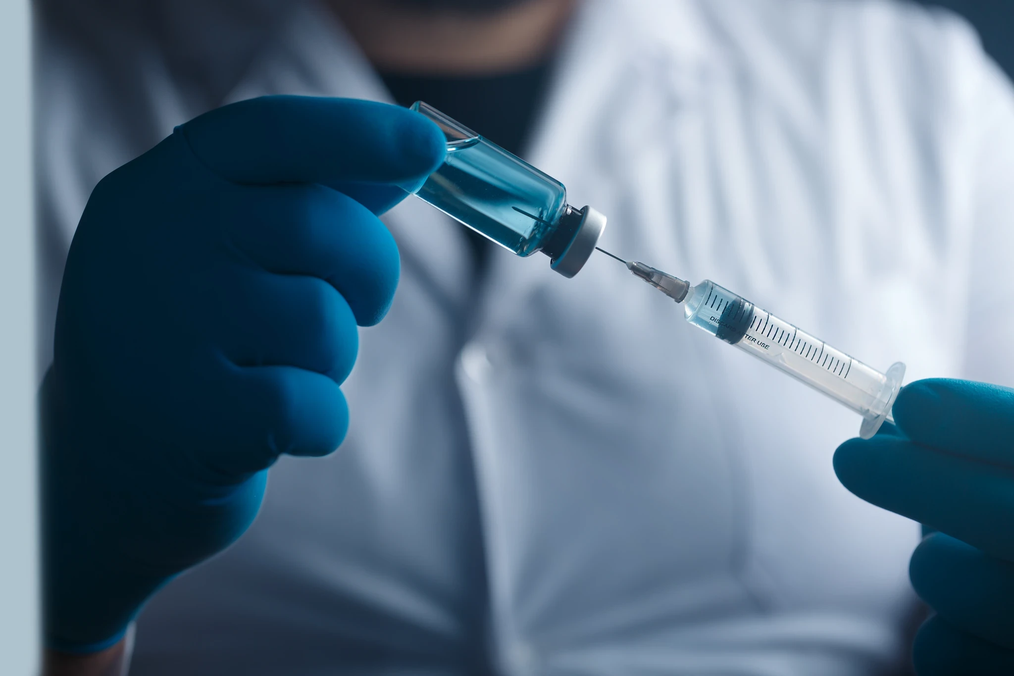 Gloved hands drawing blue liquid from a vial into a syringe.