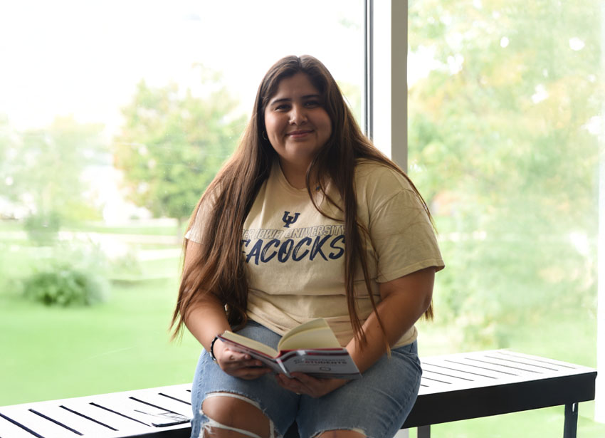 A student reads a book and smiles at the camera in front of large glass windows overlooking a green lawn