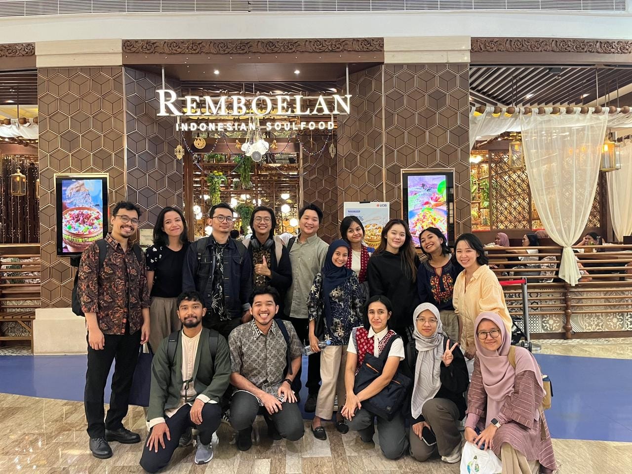 Thirteen team members standing and kneeling in front of the Remböelan Indonesian Soulfood restaurant entrance, posing as a cohesive design and research unit.