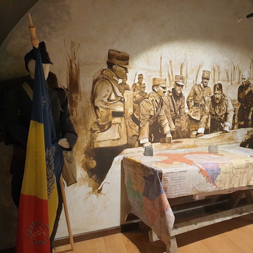 A mannequin in military uniform holds a flag next to a mural of soldiers gathered around a table covered with a map.