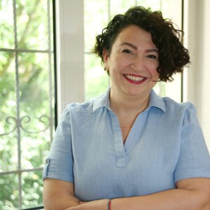 A young person with curly hair smiles at the camera while sitting in front of a laptop against a dark background.