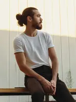 Man with long hair and beard, sitting outdoors during sunset, wearing white t-shirt and dark pants, with thoughtful expression.