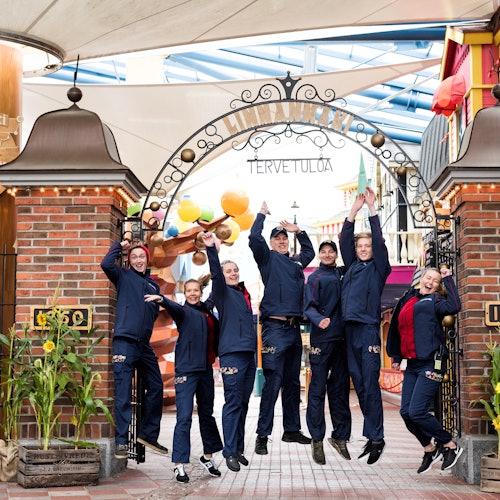 Eight people in matching navy blue uniforms jump in the air under an ornate "Tervetuloa" archway in a festive area.