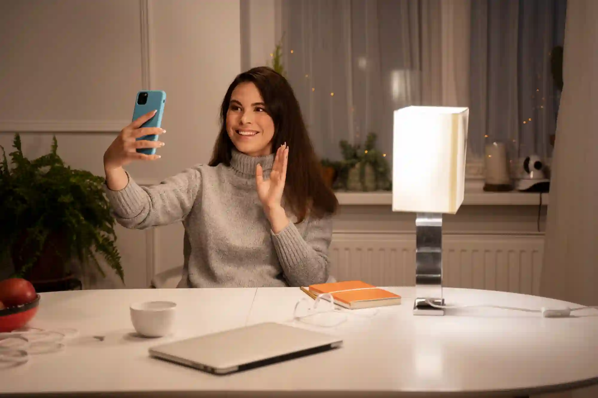 A smiling woman waves at her smartphone while filming a video or taking a selfie in a cozy room.