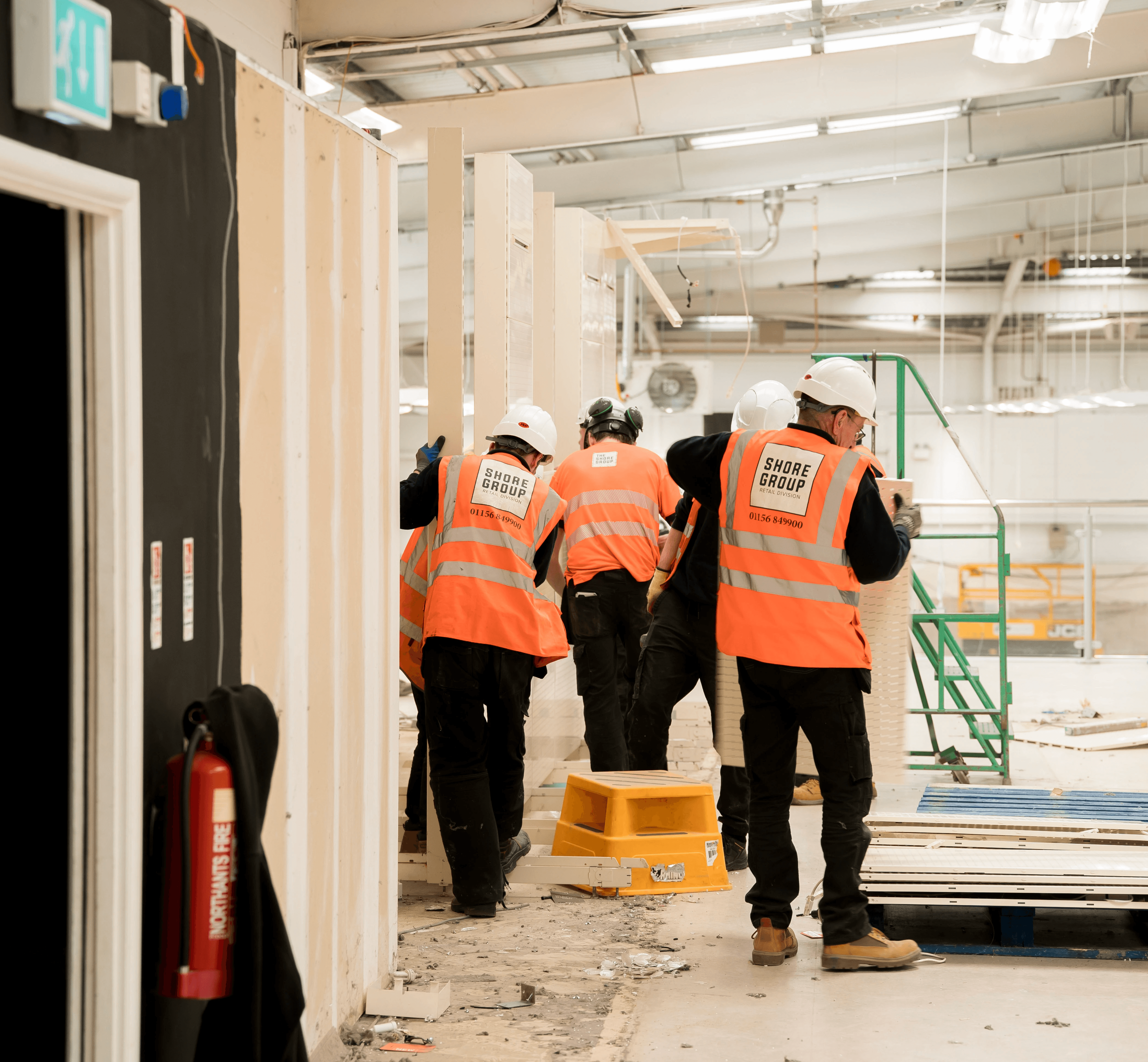 A group of construction workers wearing high-visibility jackets and hard hats celebrate at a construction site with safety signs and equipment visible in the background.