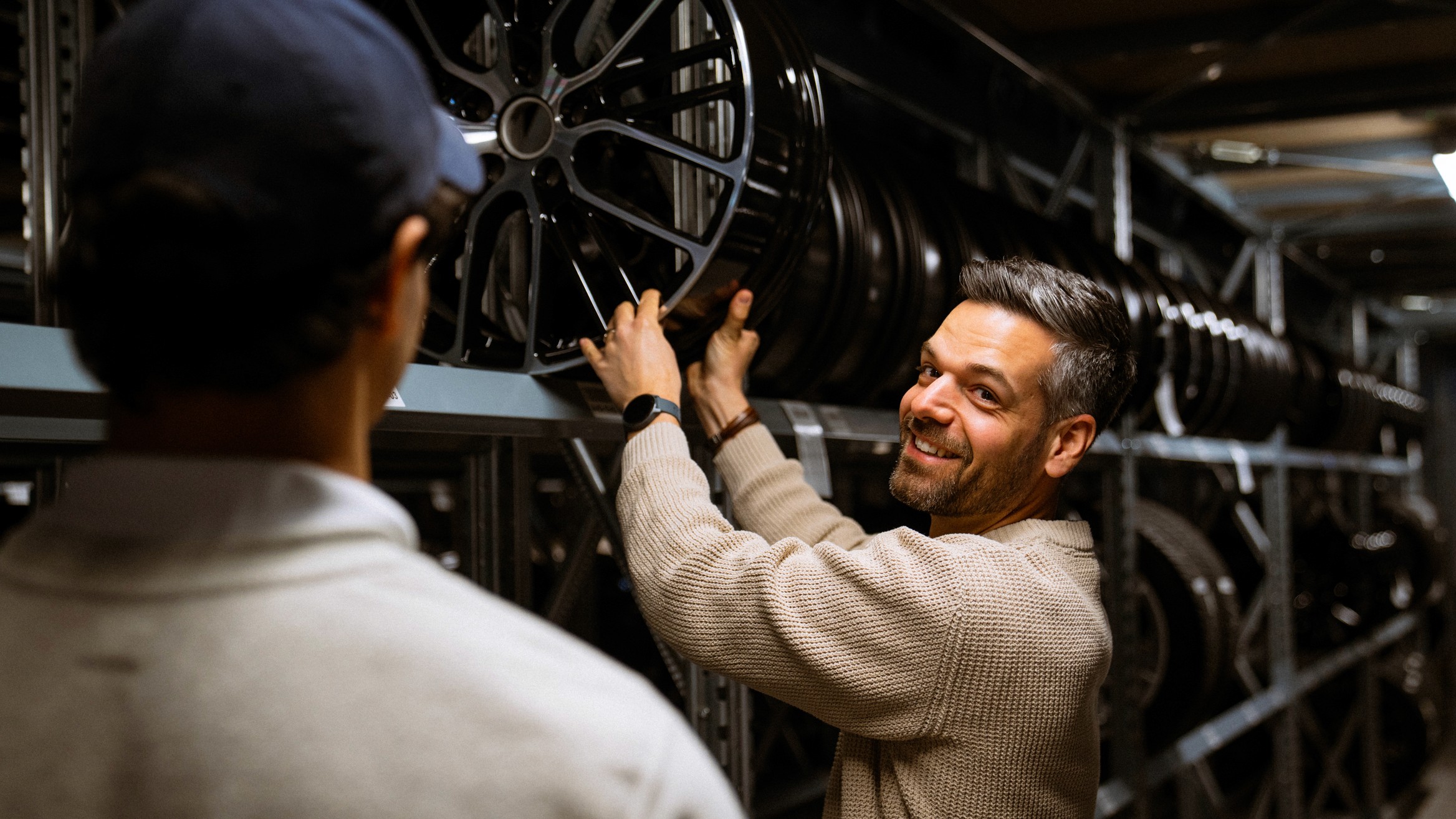 Mitarbeiter in Felgenlager nimmt eine Porsche-Felge aus dem Regal, freundliche Beratungsszene mit zweiter Person im Vordergrund