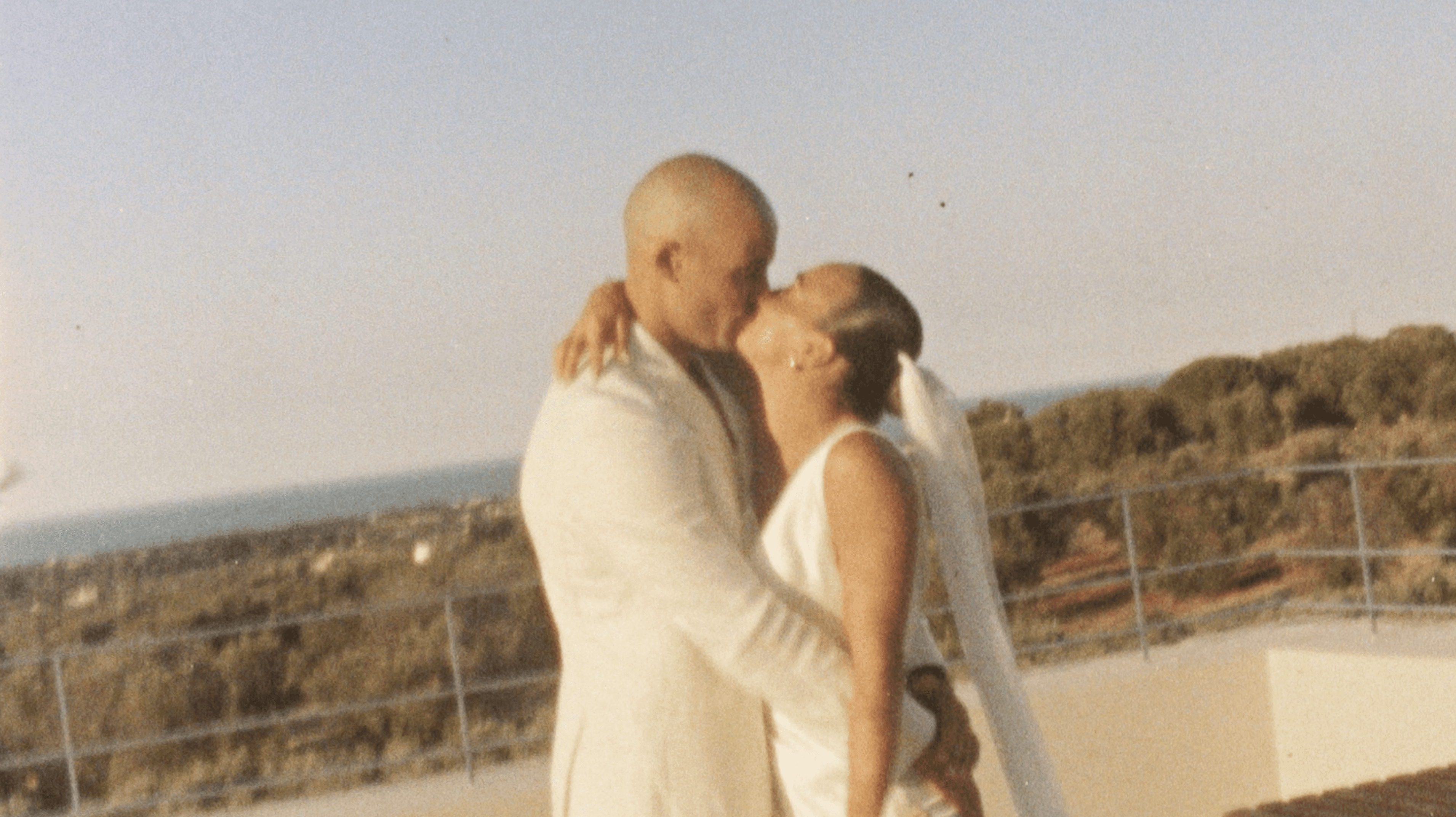 A couple in elegant white attire joyfully runs hand-in-hand outside a rustic stone building, with the bride holding a bouquet of greenery, capturing the essence of a romantic and adventurous wedding escape.