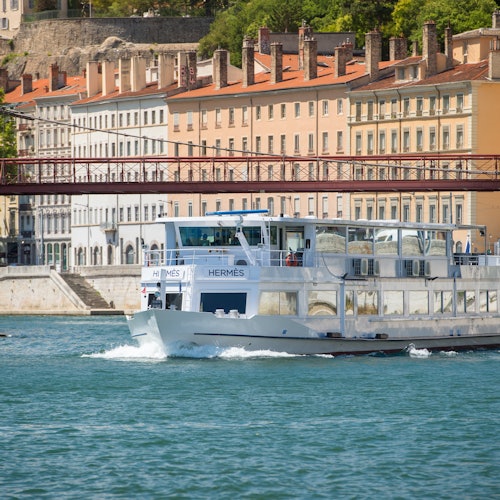 A white river cruise boat labeled "Hermès" sails on a river with historic buildings and a red bridge in the background.