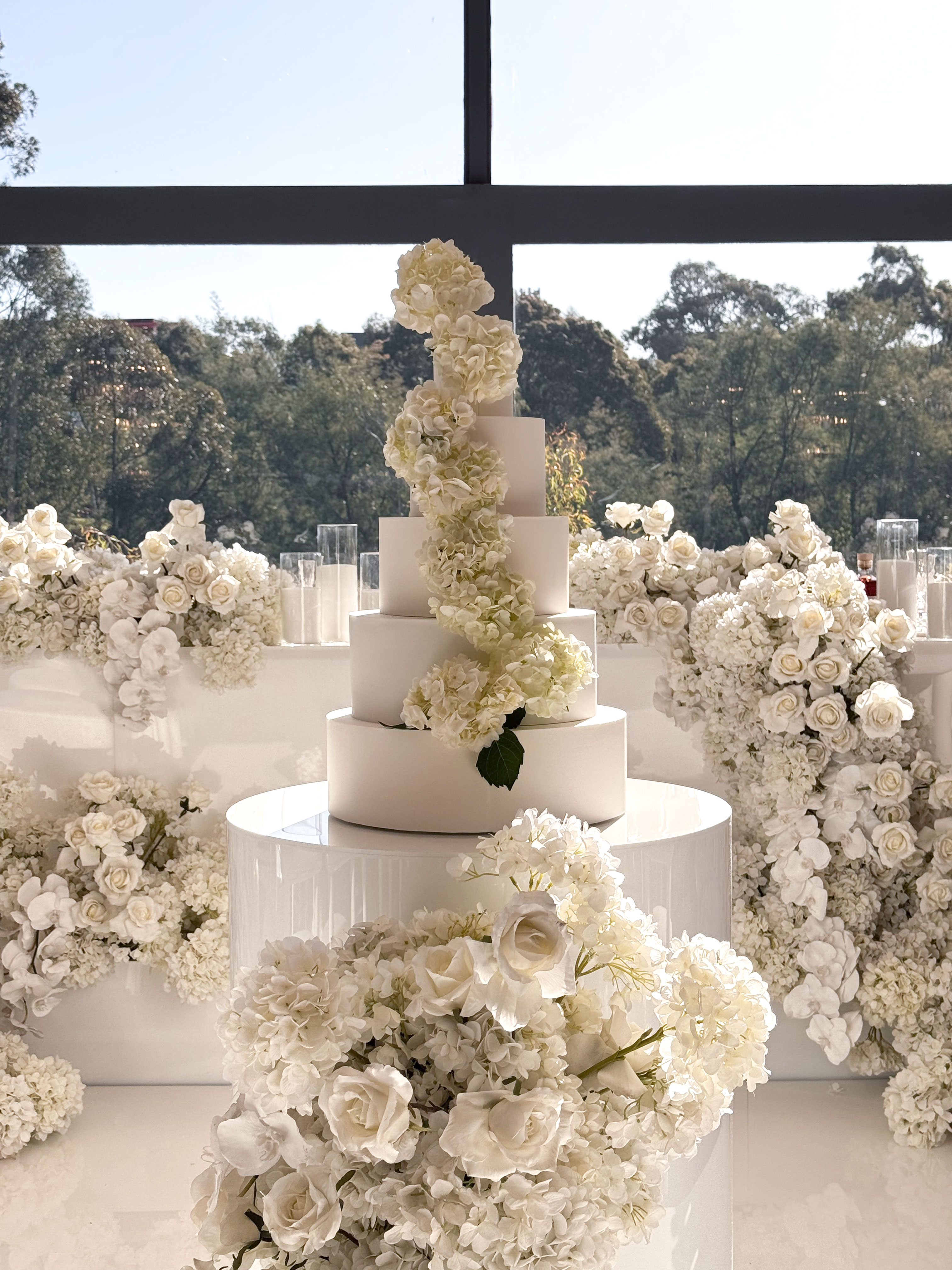 Multi tier white wedding cake with smooth fondant finish styled with cascading white hydrangeas and roses in a modern venue setting