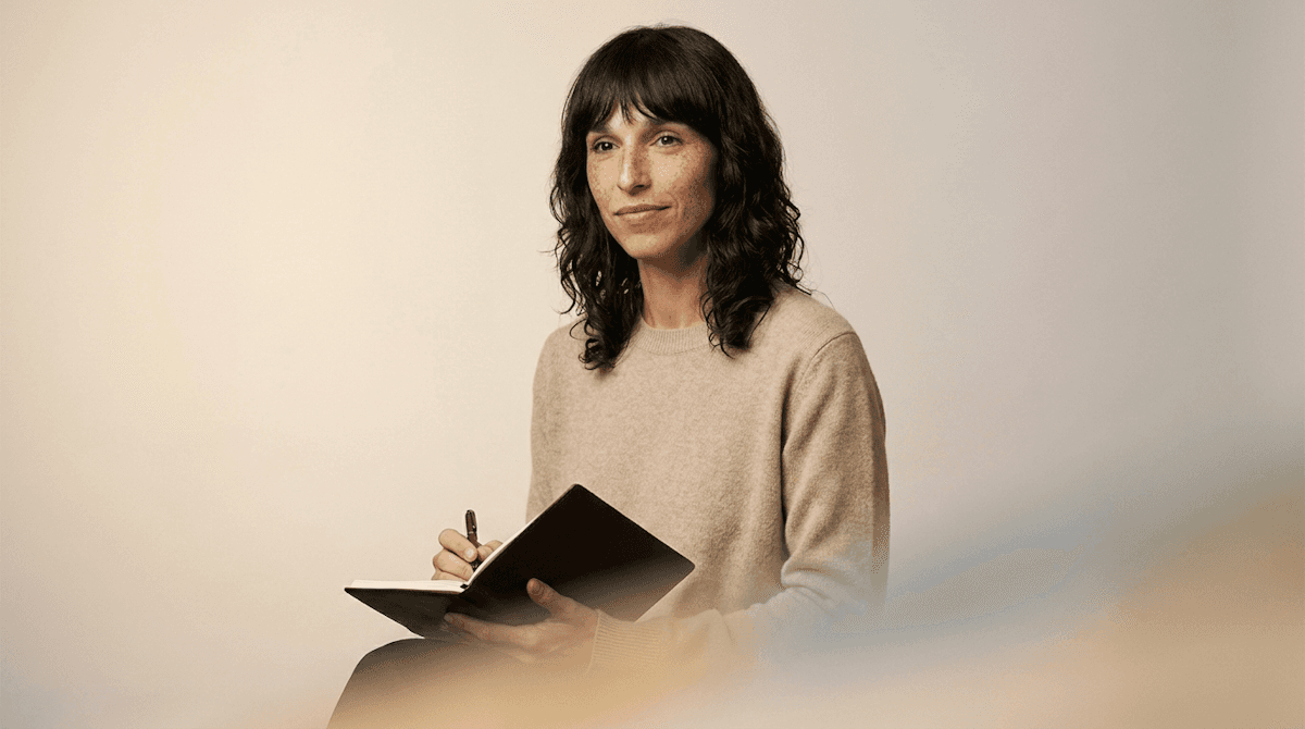 A person with dark wavy hair and a beige sweater is writing in a notebook, seated against a soft neutral background.