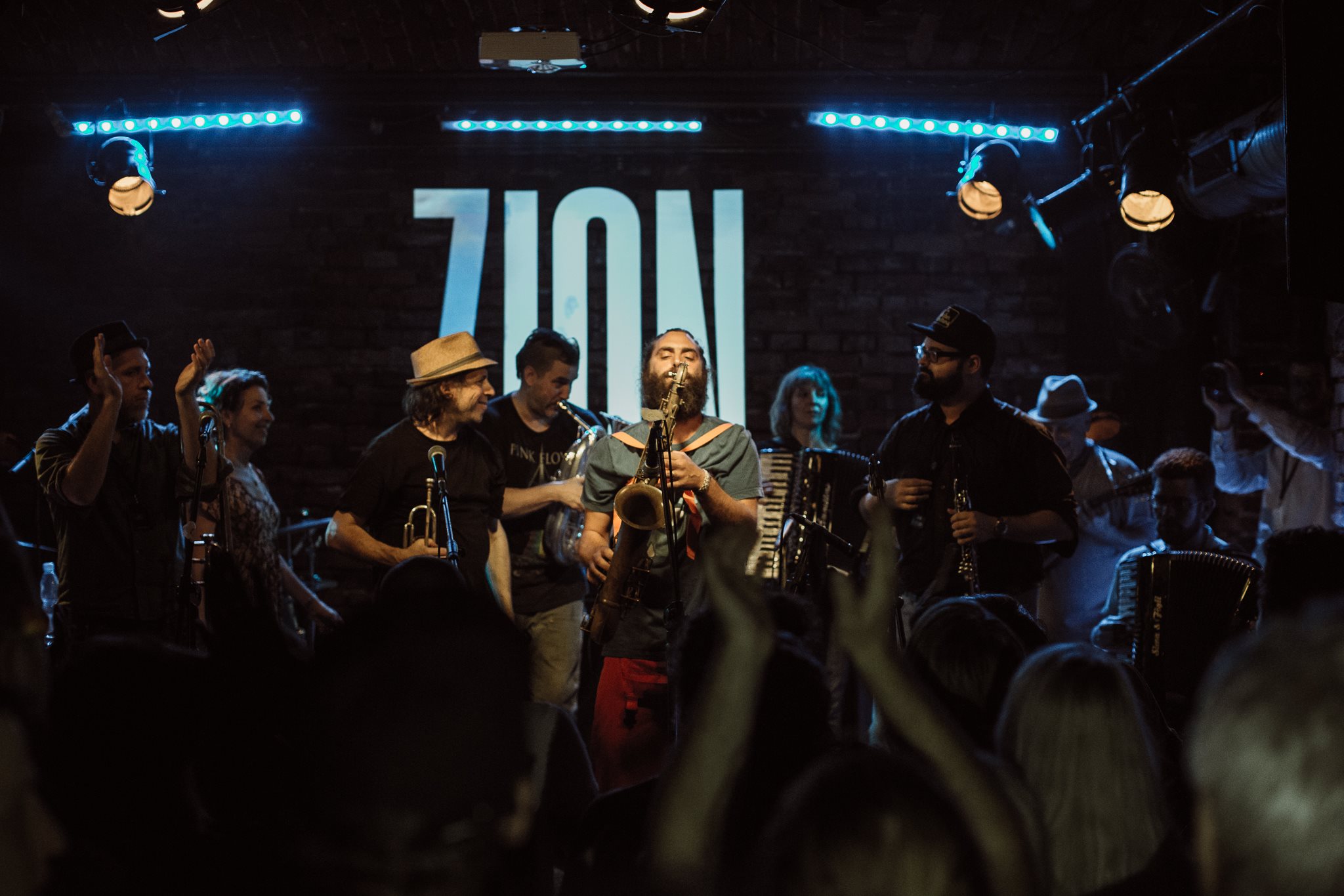 Zespół muzyków gra przed publicznością w ciemnym wnętrzu; za sceną widać duży podświetlony napis „ZION”.