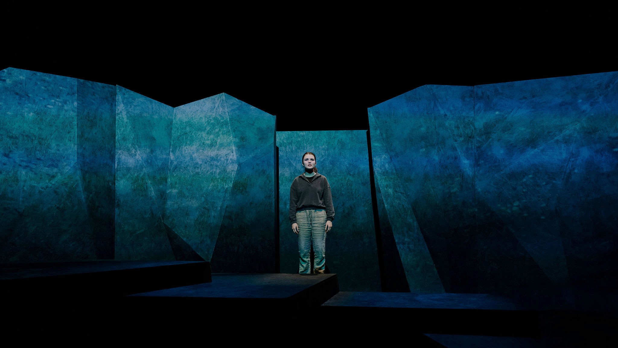 A young performer stands alone on a dark stage surrounded by jagged blue-green set pieces resembling large rock formations.