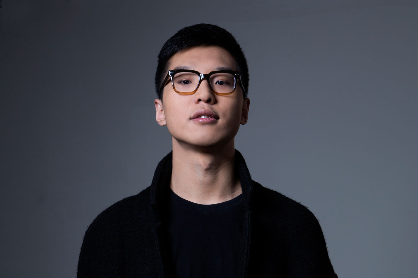 A person with short dark hair and glasses wears a black jacket over a black shirt. The background is a solid dark gray.