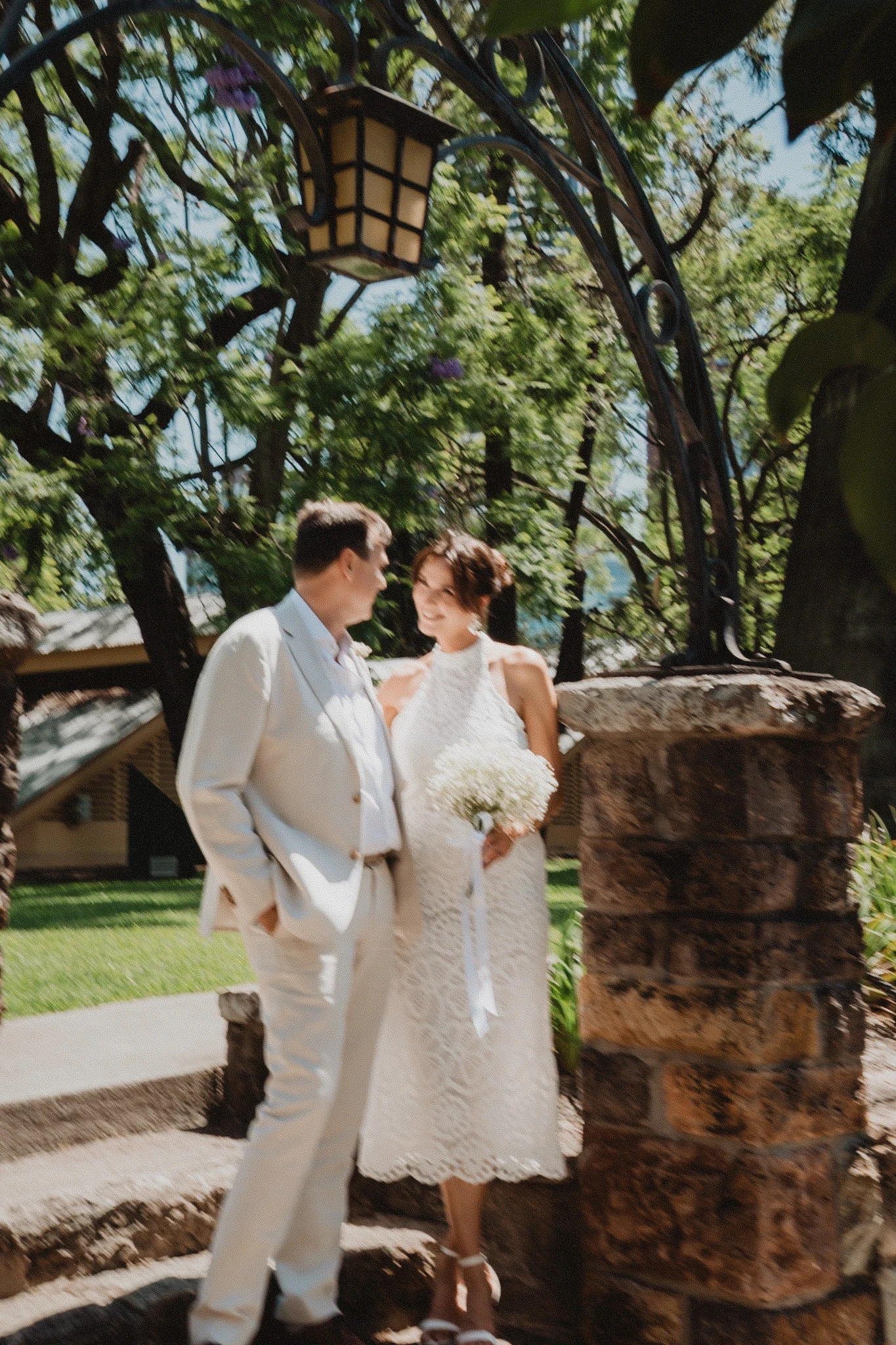 Bride and groom in white standing beneath a leafy garden arch beside a stone wall.